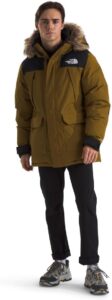 Male model wearing The North Face Men’s McMurdo Bomber Jacket in brown with faux-fur hood, paired with black pants and hiking sneakers.