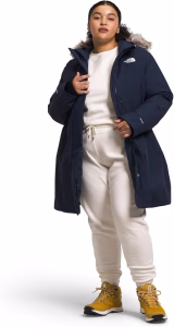 Female model wearing The North Face Women’s Arctic Parka in navy blue with faux-fur hood, styled with cream joggers and yellow hiking boots.